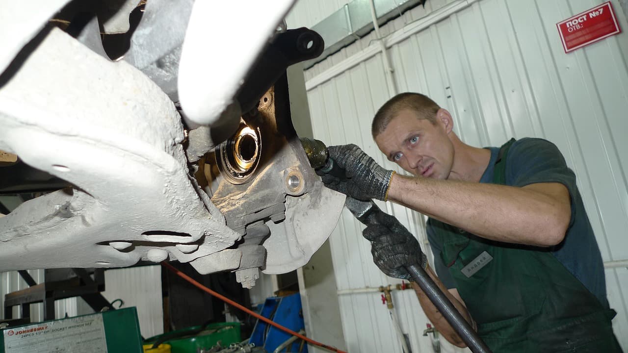 A mechanic in gloves working on a car's front wheel hub from underneath